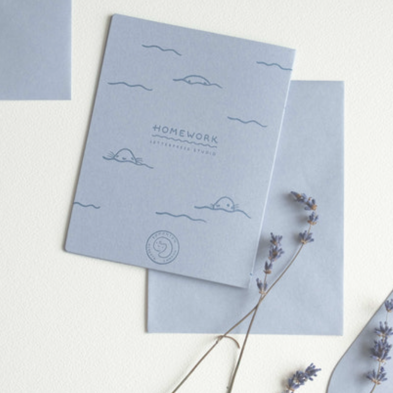 A light blue stationery card or notebook cover with simple cloud illustrations and the text 'HOMEWORK LETTERPRESS STUDIO,' layered over a matching blue sheet on a white background with dried lavender sprigs.