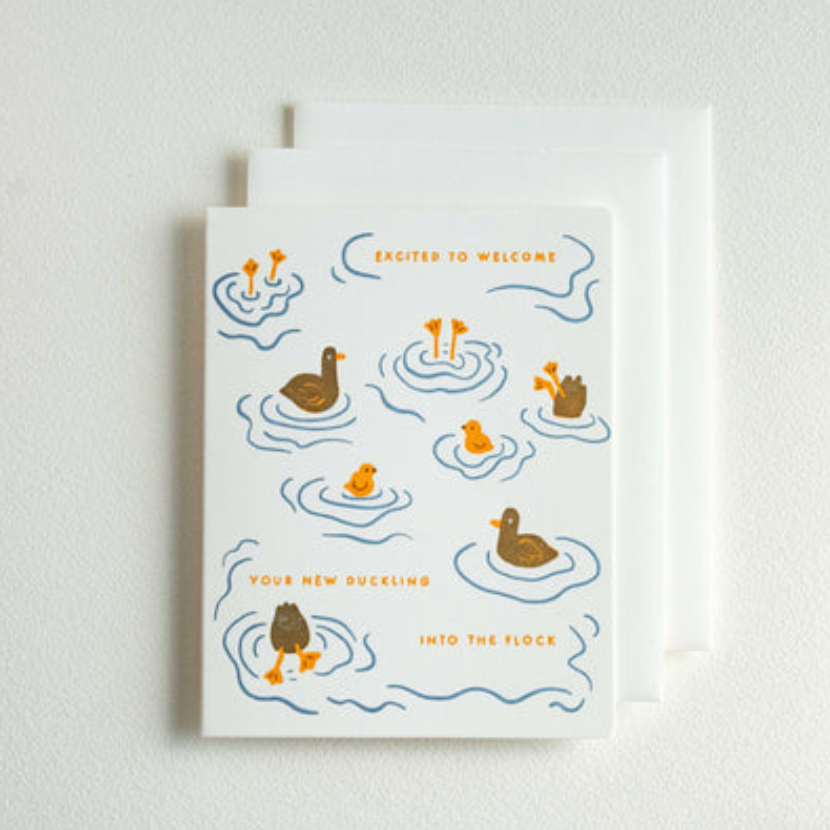 A white greeting card with a duck-themed illustration and text reading 'Excited to Welcome' and 'Our New Duckling Into the flock,' shown with a white envelope behind it on a light wall.