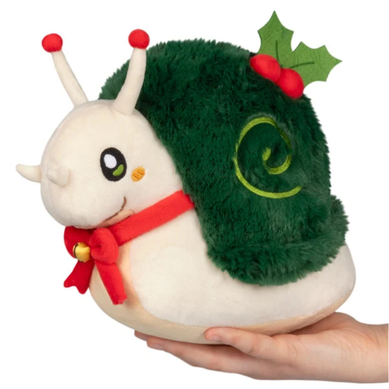 A plush snail toy with a cream-colored body, a fluffy dark green shell decorated with a green spiral and holly leaves, and a red bow and red antennae, being held in a person's hand against a white background.