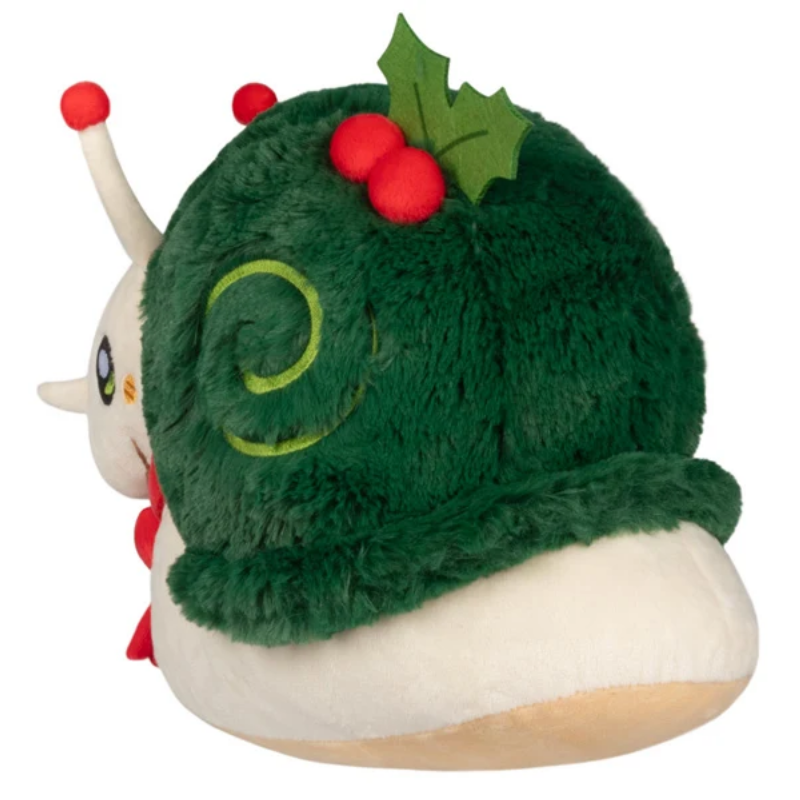 A plush toy shaped like a snail wearing a green Christmas hat with red berries and holly leaves, on a plain white background.