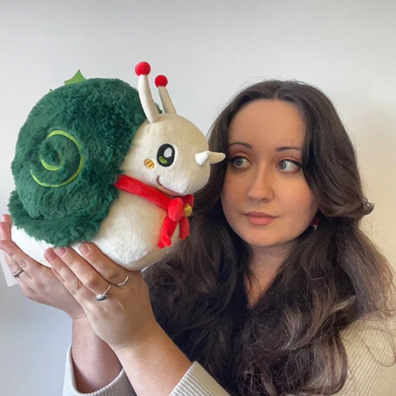 A woman holding a plush toy snail with a green shell, cream-colored body, and a red scarf against a plain light gray wall.