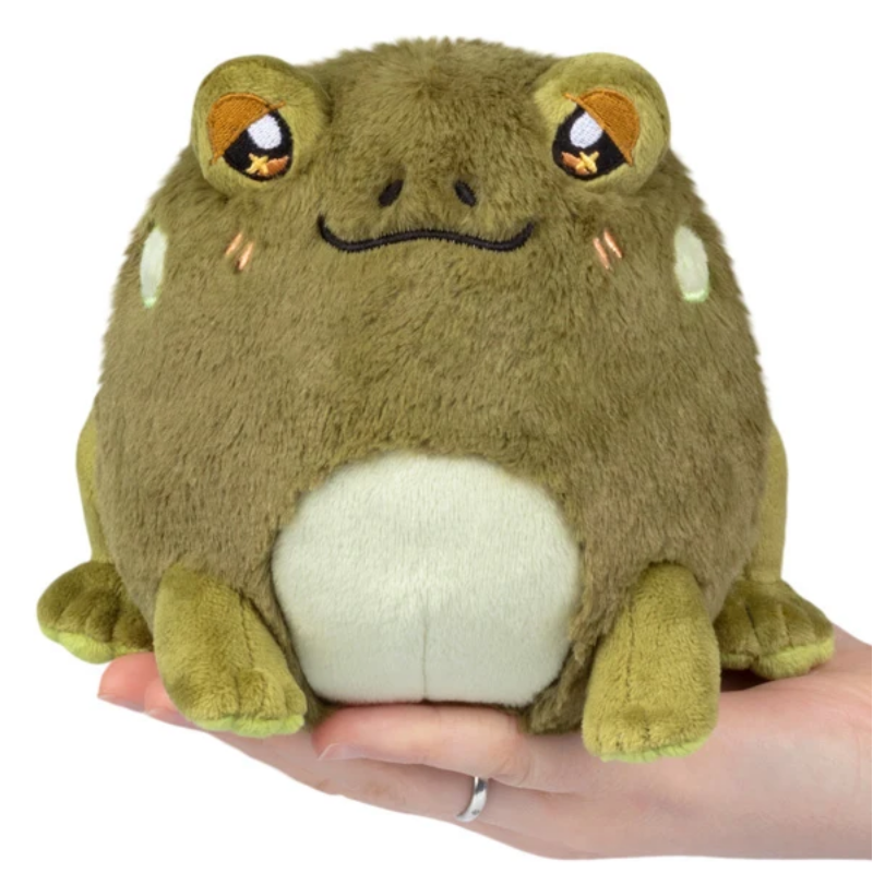 A plush frog toy with olive green fur, a cream-colored belly, and embroidered orange eyes, being held in a person's hand against a white background.