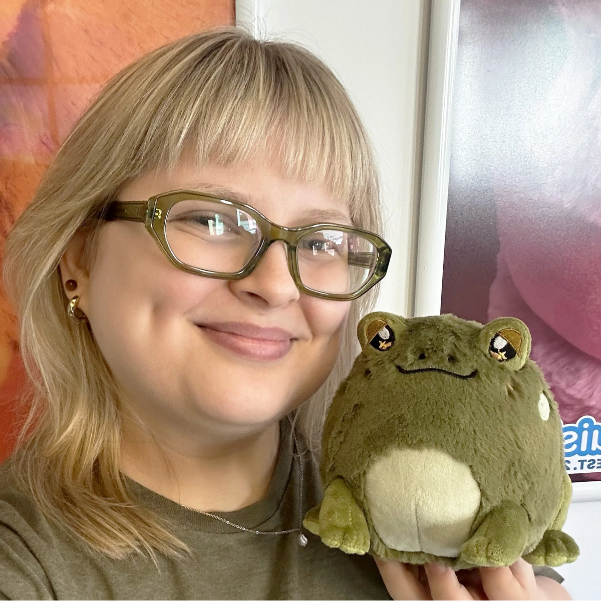 A smiling woman with blonde hair and glasses holds a plush green frog toy with a cream belly in front of an indoor background with posters.
