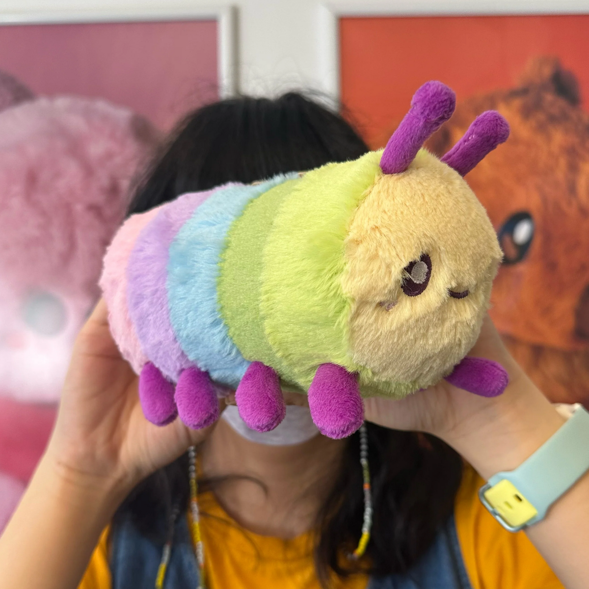 A person holds a plush caterpillar toy in front of their face. The caterpillar has pastel rainbow stripes in purple, blue, green, yellow, and cream, with purple antennae and legs.