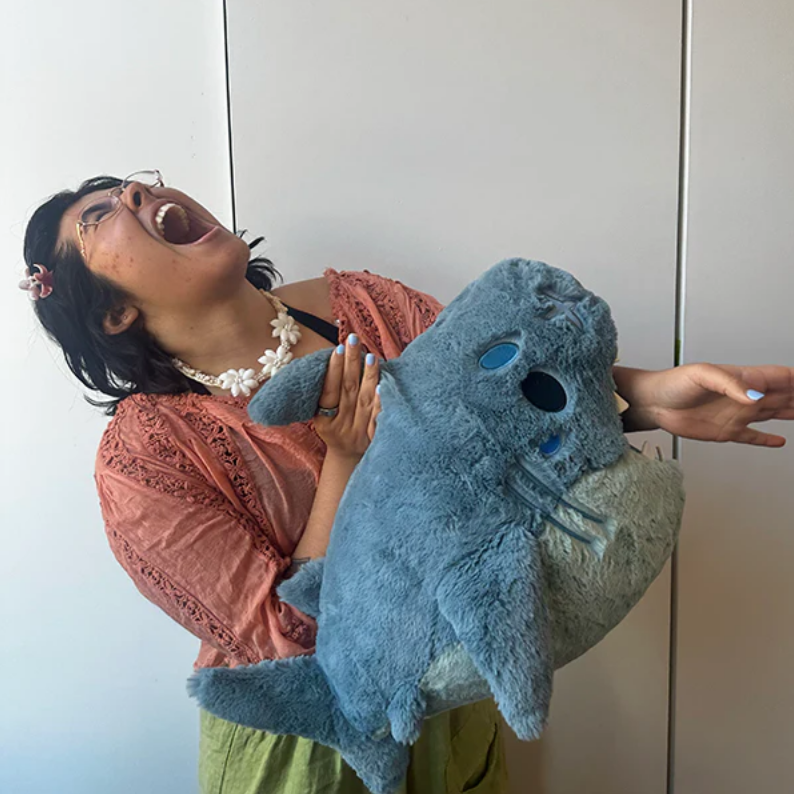 A person in a peach lace top and green skirt holds a large blue plush sea creature toy with blue eyes and a white underside against a plain indoor wall.