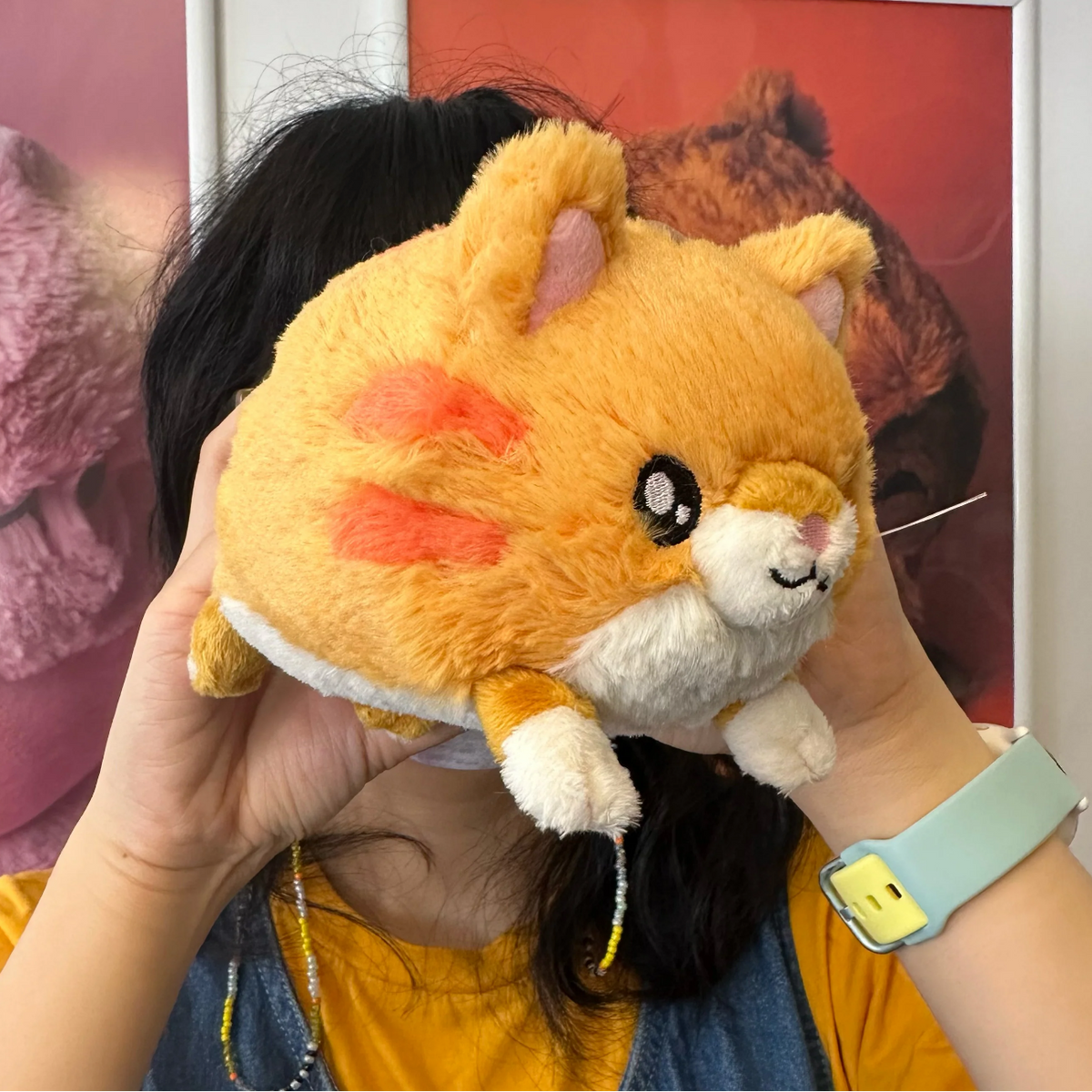 A person holds a round orange plush toy with a cat-like face and pink ear details in front of their face. The person is wearing a yellow top and a light blue smartwatch band, with posters in the background.