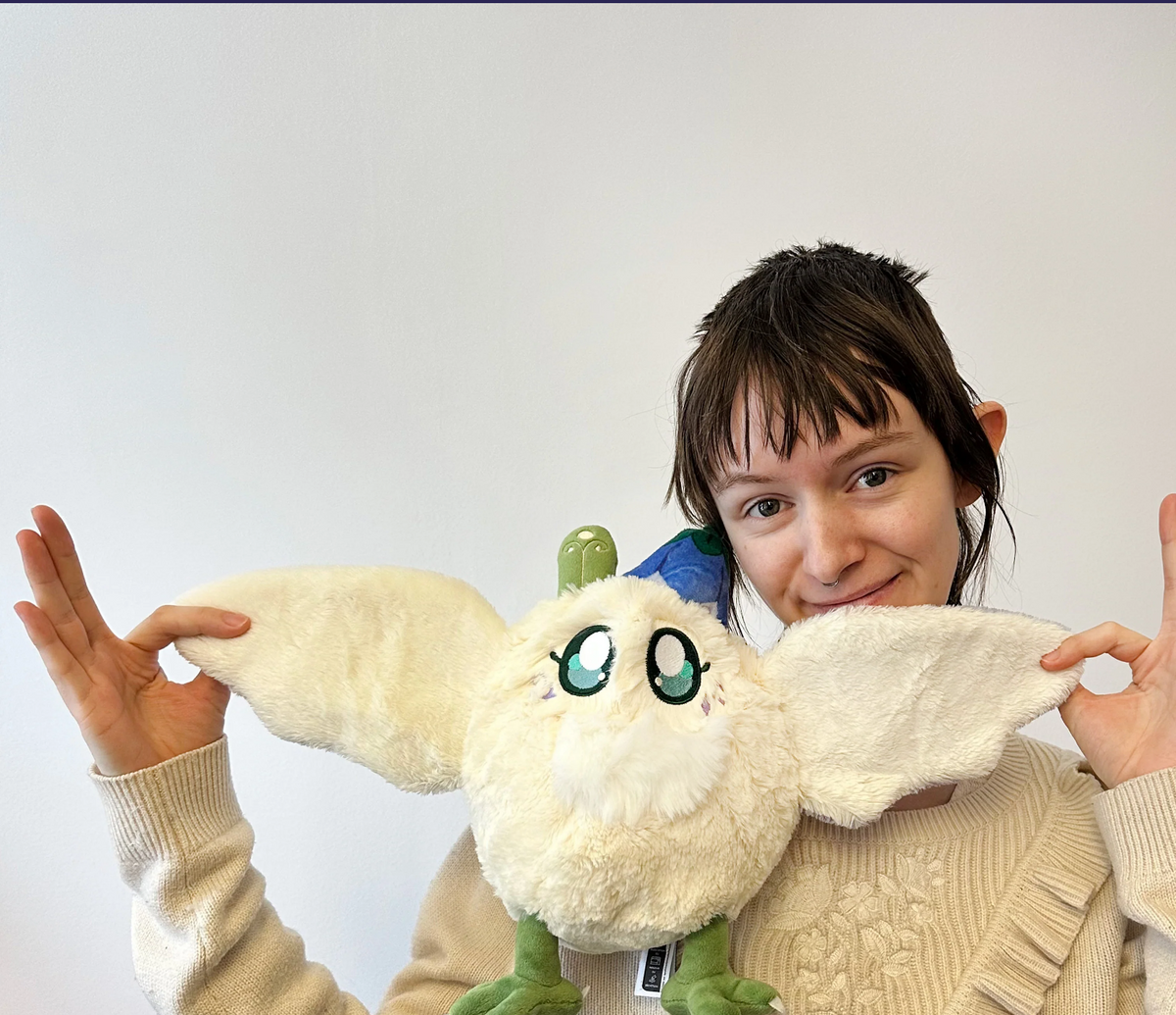 A person holding a plush toy with large cream-colored wings and green eyes, posing in front of a plain light gray wall.