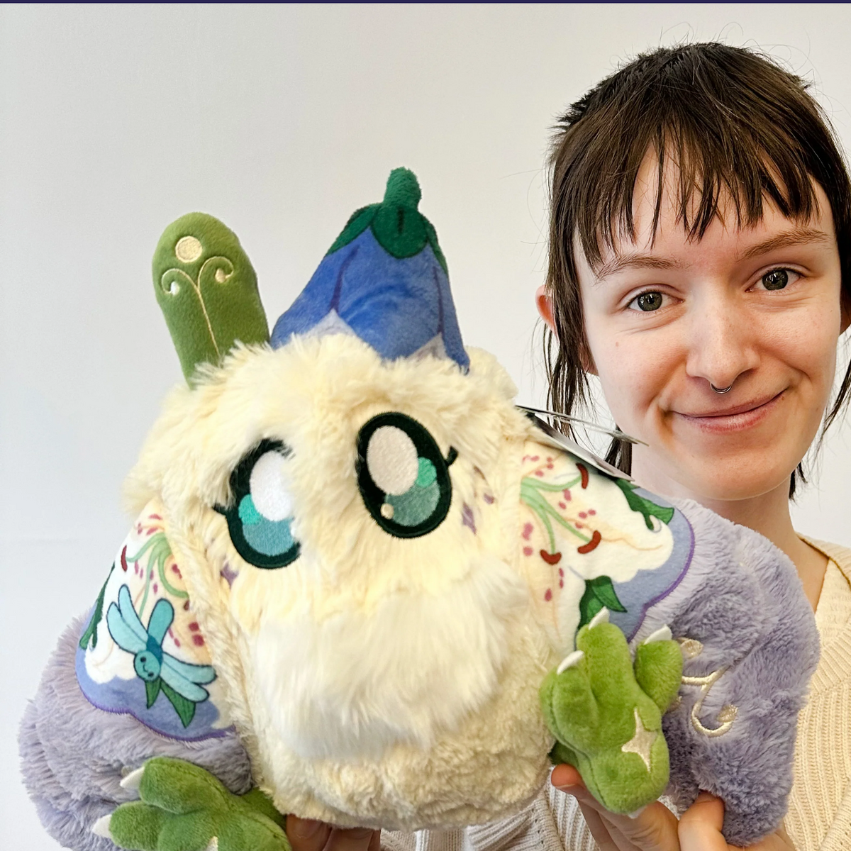A smiling woman holds a fluffy plush toy with large embroidered eyes, green limbs, and pastel blue and purple accents against a plain light background.
