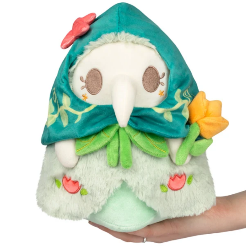 A plush hooded character toy being held in a person's hand against a plain white background. The hooded figure has a teal-green hood with embroidered floral details, a long white snout, brown eyes, and a pale mint-green body with small pink and orange flower accents.