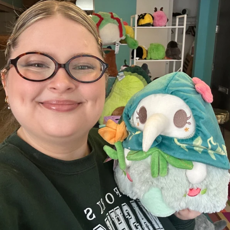 Person holding a plush gnome toy with a colorful hat in a room with shelves of more plush toys.