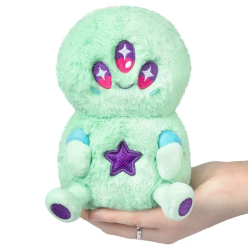 A mint green plush toy with purple and pink embroidered eyes, a purple star on its chest, and light blue accents on its arms and feet, being held in a person's hand against a white background.