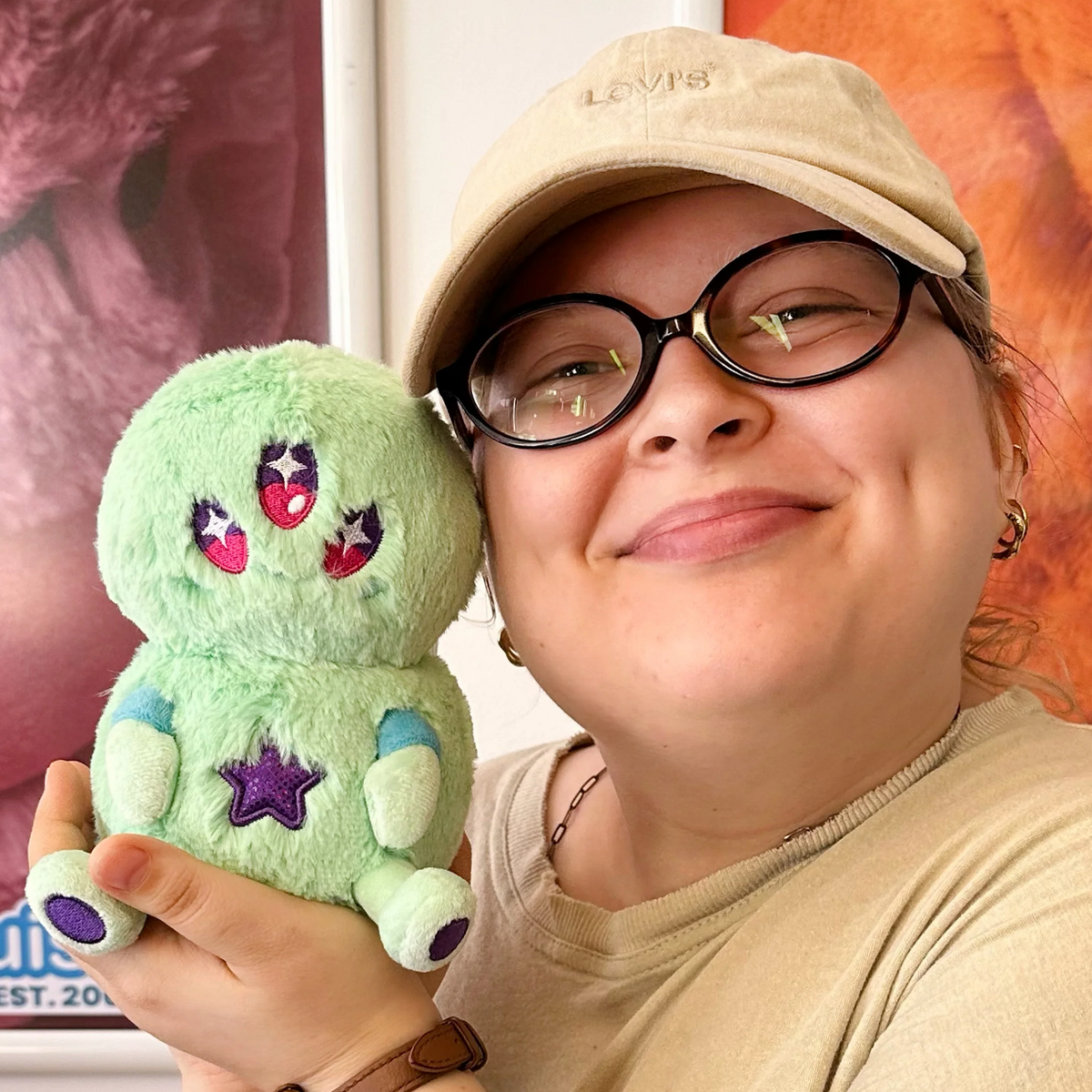 A person wearing a beige cap with 'LOVIS' embroidered on it holds a light green plush toy with embroidered pink and purple details. The background shows a colorful framed print.