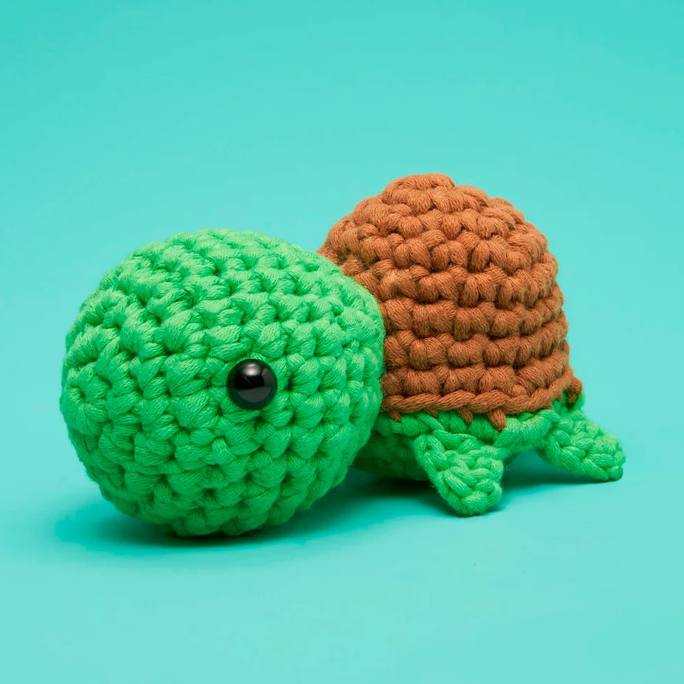 A crocheted turtle toy with a bright green head, flippers, and back, and a brown shell, photographed on a smooth turquoise background.