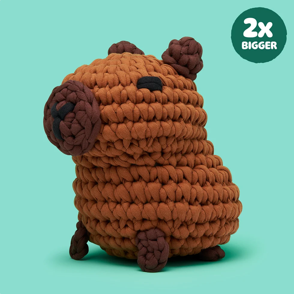 A brown crocheted capybara plush toy with dark brown accents, shown on a light teal background with a green circular badge in the top right that reads '2X BIGGER'.