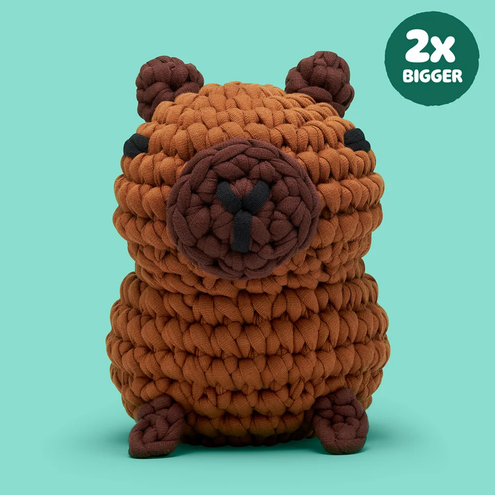 A plush capybara toy made from chunky brown yarn, shown against a solid mint green background with a dark green circular badge in the top right that reads '2x BIGGER'.