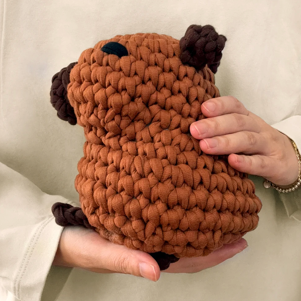 A handmade, chunky knit brown plush toy with dark brown ears and nose, held in two hands against a soft beige background.