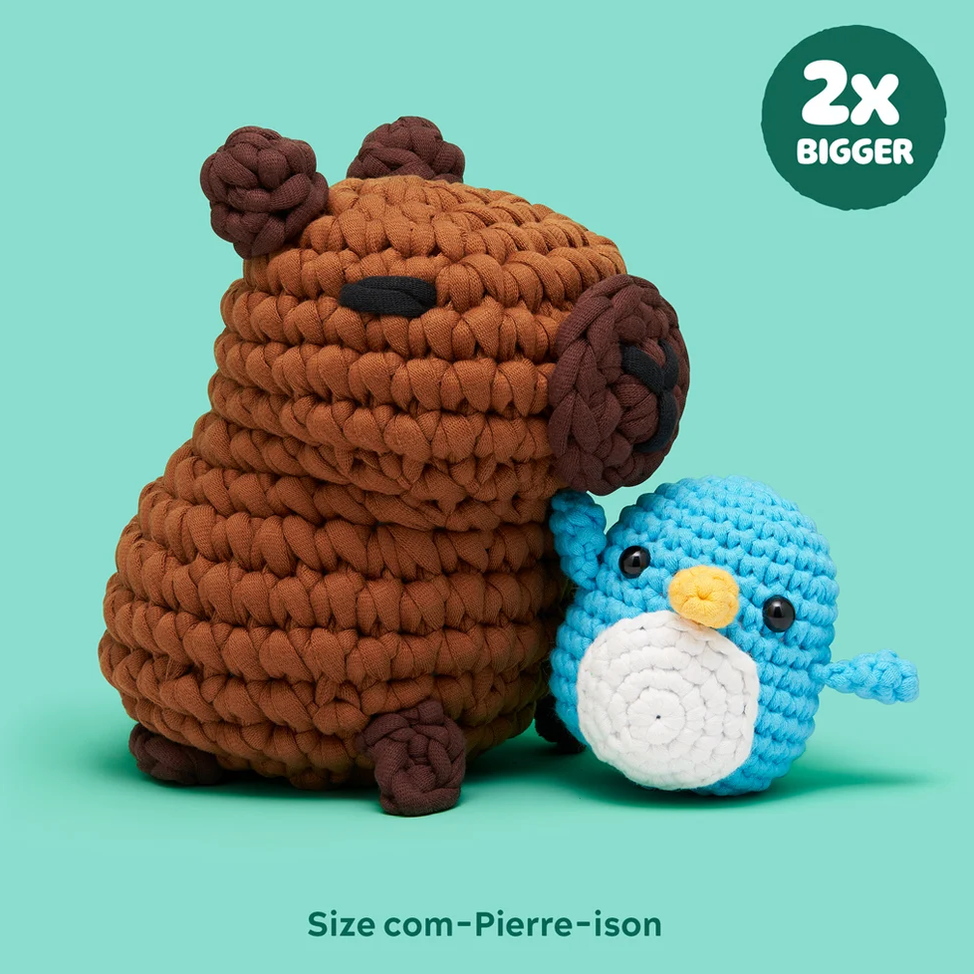 Two crocheted capybara plush toys on a mint green background, with one large brown capybara and one smaller blue capybara. A dark green circular badge in the top right says '2x BIGGER', and text at the bottom reads 'Size com-Pierre-ison'.
