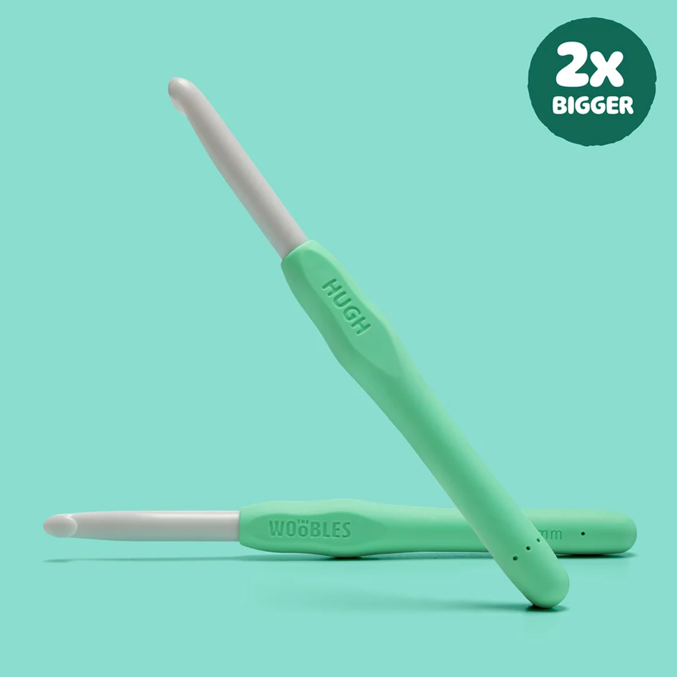 Two mint green crochet hooks with white tips, one standing upright and one lying horizontally, on a light aqua background. A dark green circular badge in the top right reads '2x BIGGER'.