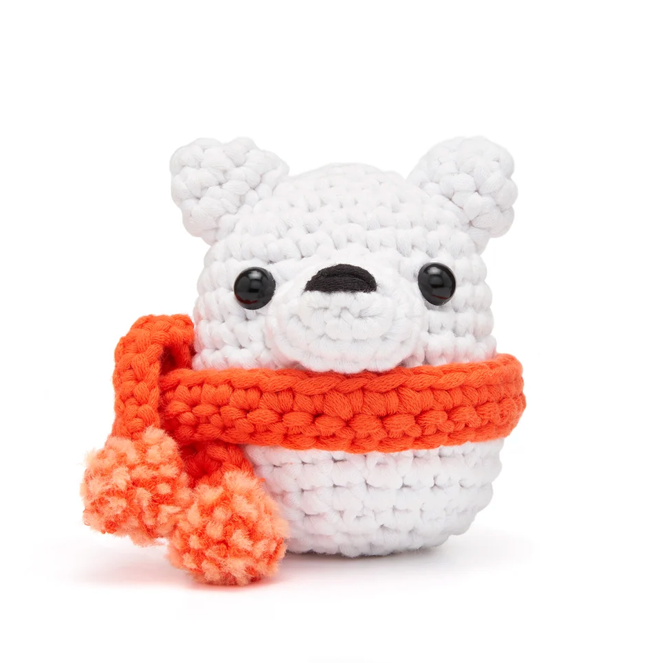 A white crocheted polar bear plush toy wearing an orange scarf, featuring black bead eyes and a black nose, set against a clean white background.