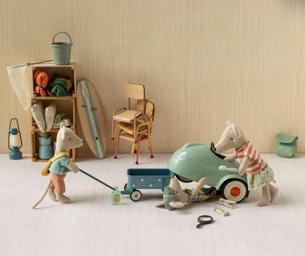 A styled indoor play scene with wooden animal figurines and toy accessories. A mouse pushes a blue wagon while a horse and cow sit in a mint green toy car, with shelves, chairs, a surfboard, and other beach-themed toys arranged against a beige wall.