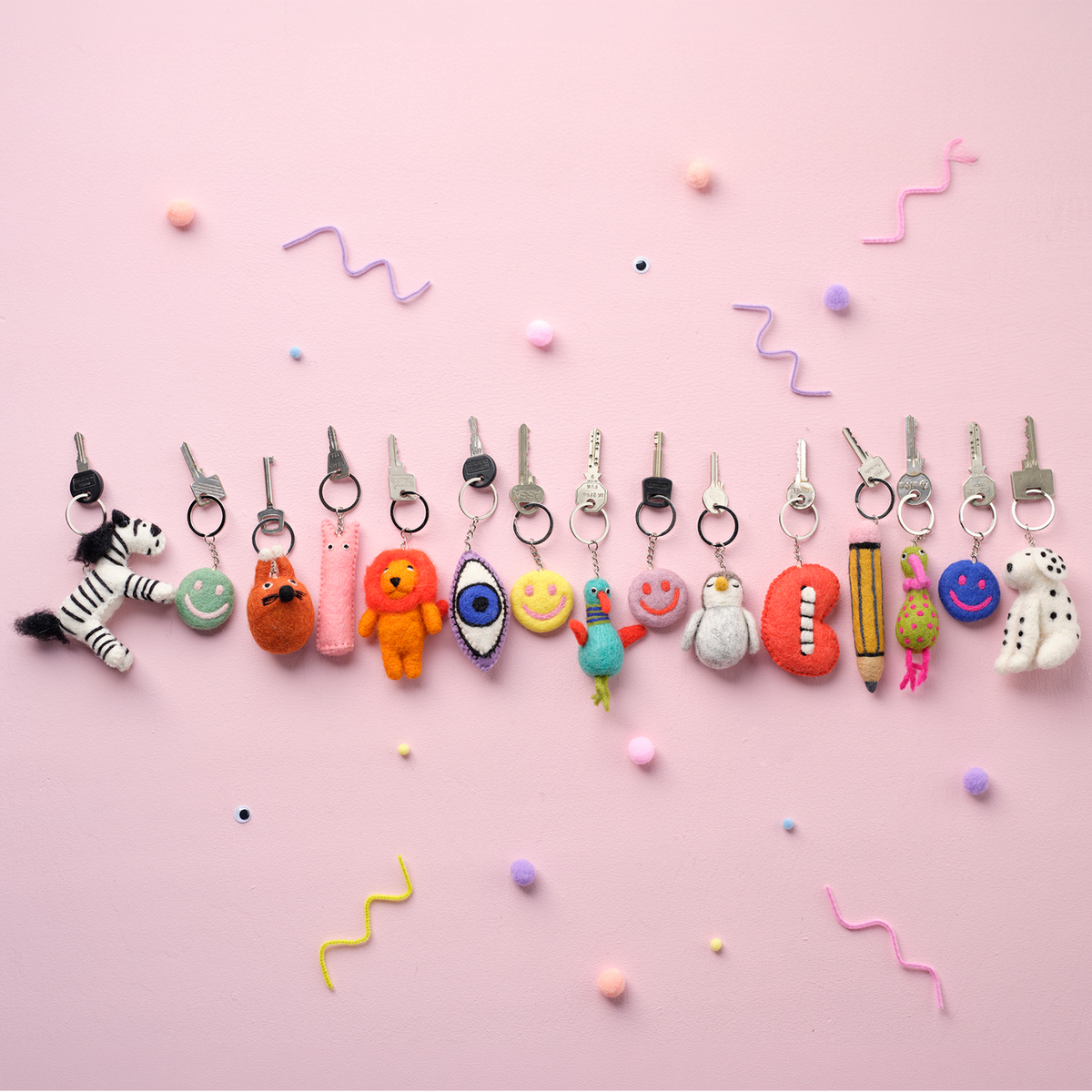 A row of colorful novelty keychains attached to metal keys, arranged horizontally on a light pink background with small pastel decorative dots and squiggle marks.