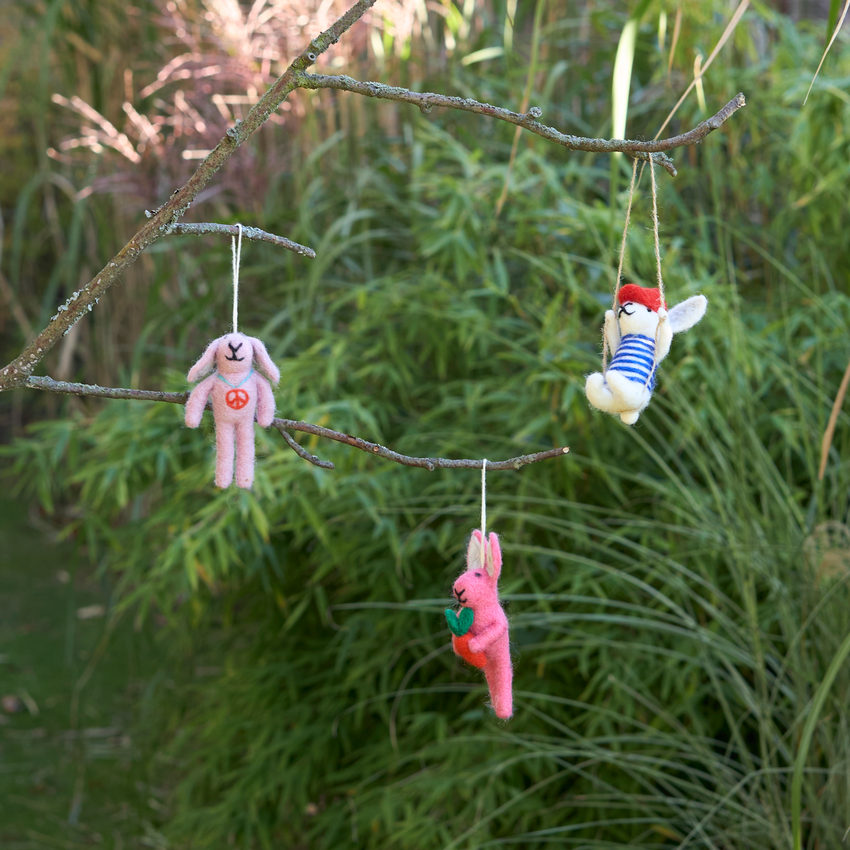 Three small handmade plush bunny toys in pink, white, and red are hanging from thin strings attached to a tree branch in an outdoor garden setting with green reeds in the background.