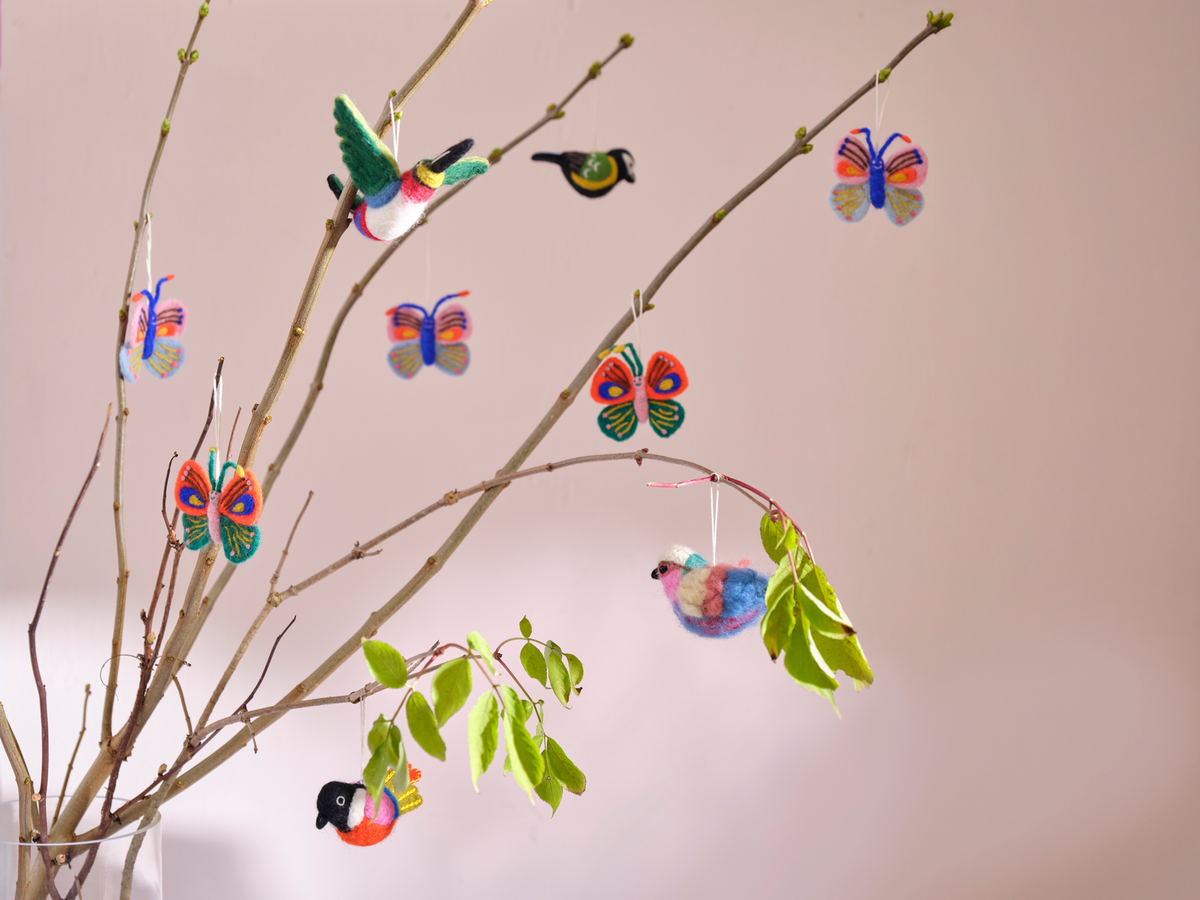 Handmade colorful felt bird and butterfly ornaments hanging from thin branches with small green leaves against a soft pink background.