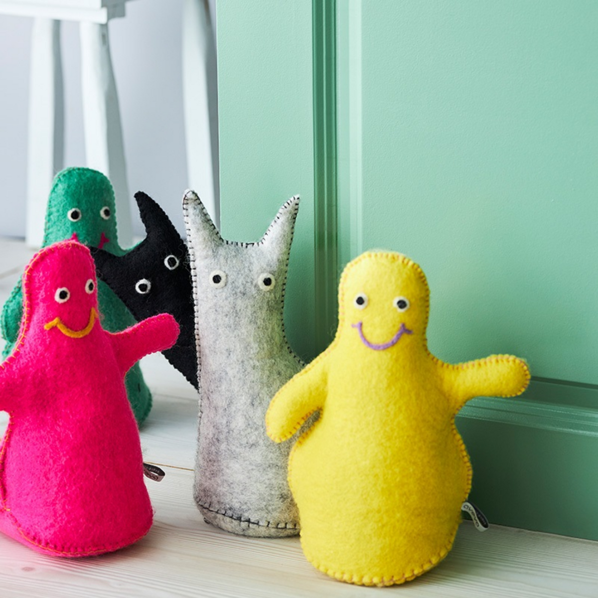 Four fuzzy monster-shaped plush toys in bright pink, yellow, black, and gray, arranged on a light wood surface against a mint green wall.