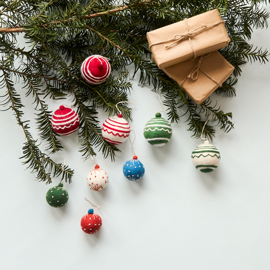 A styled flat-lay of Christmas ornaments hanging from green pine branches, with two wrapped kraft-paper gift boxes on a light gray background.
