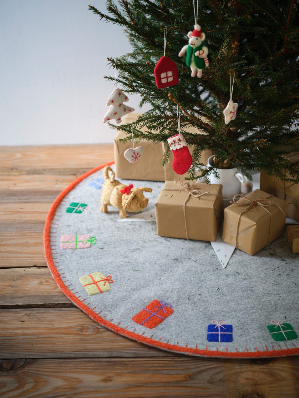 A round felt Christmas tree skirt with an orange border and colorful gift patterns, displayed under a decorated Christmas tree with wrapped presents and ornaments on a wooden floor.