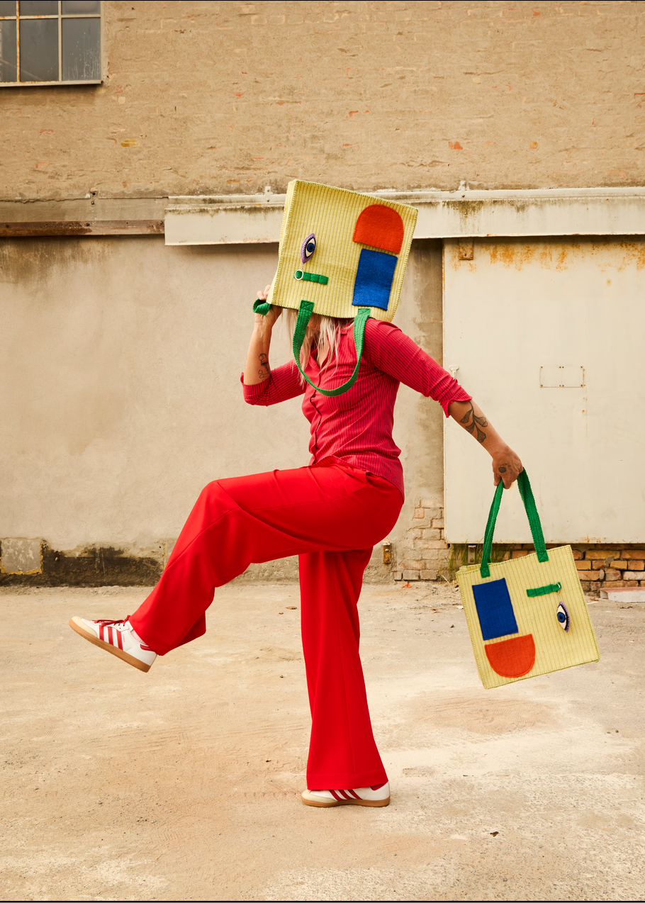 A person in a red tracksuit holds a large woven tote bag over their head and carries a smaller matching tote bag in one hand, posed outdoors against a beige concrete wall.