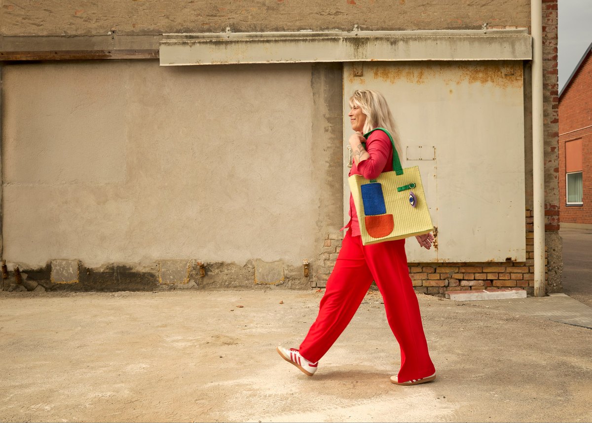 A woman in a bright pink and red tracksuit walks past a concrete wall carrying a large, colorful geometric-patterned tote bag.