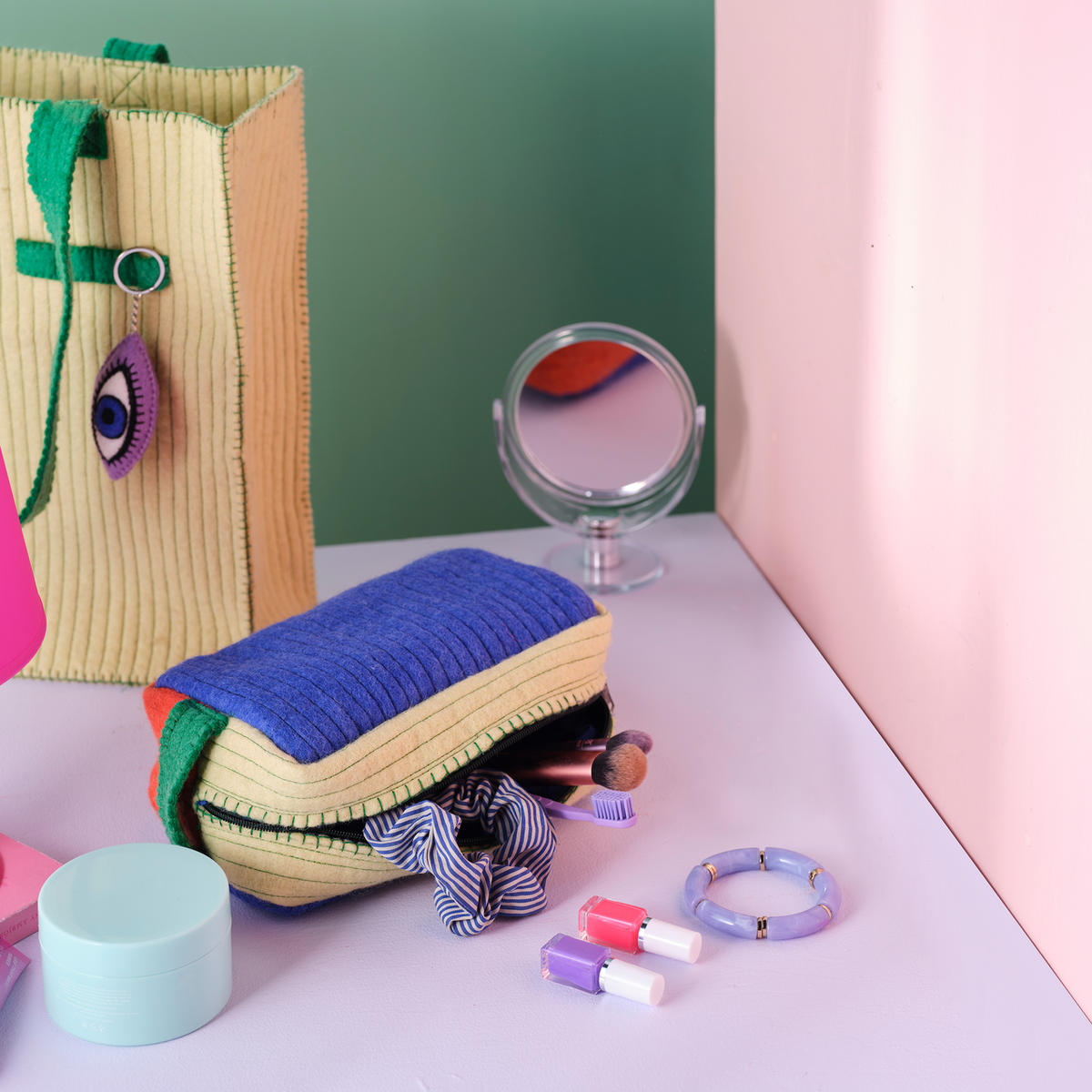 A styled flat-lay of handmade knitted cosmetic pouches and accessories on a pastel pink and green set. The main product is a striped cream and blue zippered pouch with green handles, shown alongside a matching tote bag, a small round mirror, and colorful makeup items.