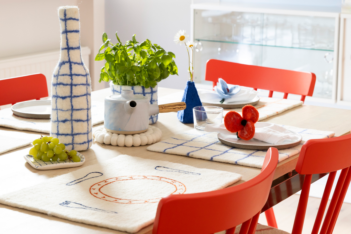A styled dining table scene with cream placemats featuring blue and orange embroidered patterns, a white teapot, a blue vase with daisies, green grapes, red flowers, and bright red chairs in a sunlit room.