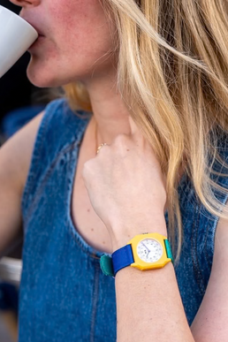A close-up lifestyle shot of a woman wearing a colorful watch with a yellow case and a blue strap, drinking from a white cup.