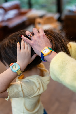 Two people wearing colorful watches, one with a child's hand on their head, in a blurred indoor setting.
