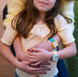 A close-up lifestyle shot of a young girl wearing two colorful smartwatches, one on each wrist, while being held by an adult. The girl is wearing a pale yellow top, and the watches feature bright yellow, blue, and orange colors.