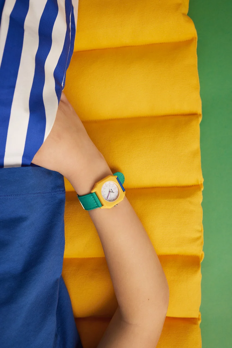 A close-up of a child's arm wearing a colorful toy watch with a yellow case, white face, and green strap, set against a bright yellow padded background and blue striped shirt.