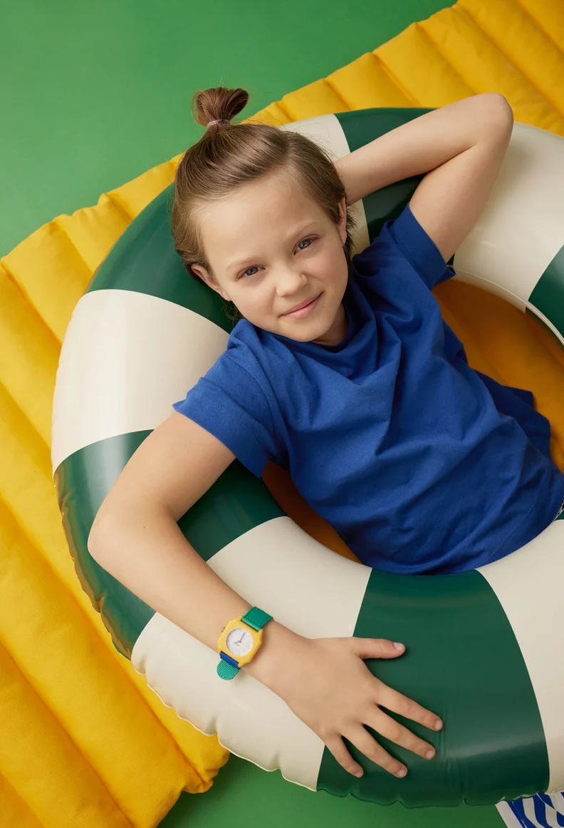A young girl with her hair in a bun lies on a green and white striped inflatable pool float, wearing a colorful smartwatch with a yellow strap and green band. She is wearing a blue t-shirt against a solid green background with a yellow inflatable mat.