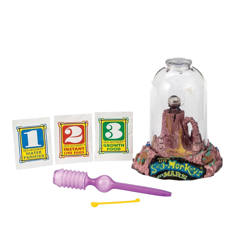 The image shows a Sea-Monkey On Mars kit which includes a plastic aquarium with a Mars landscape design at the bottom, a packet of sea monkey eggs, a water purifier, and various other accessories laid out around it.