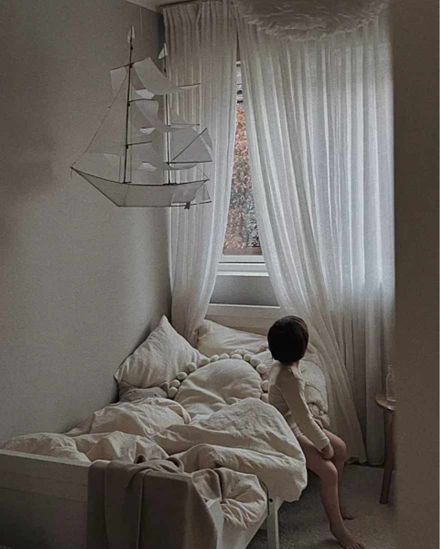 Child sitting on a bed in a softly lit room with white curtains and a ship-shaped lamp.