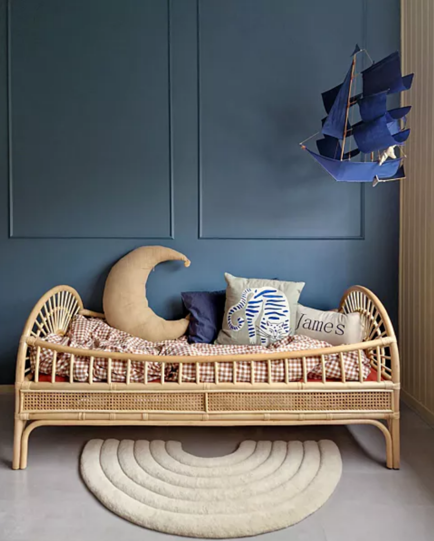 Children's bedroom with a wicker bed, decorative pillows, and a ship wall decoration against a blue wall.
