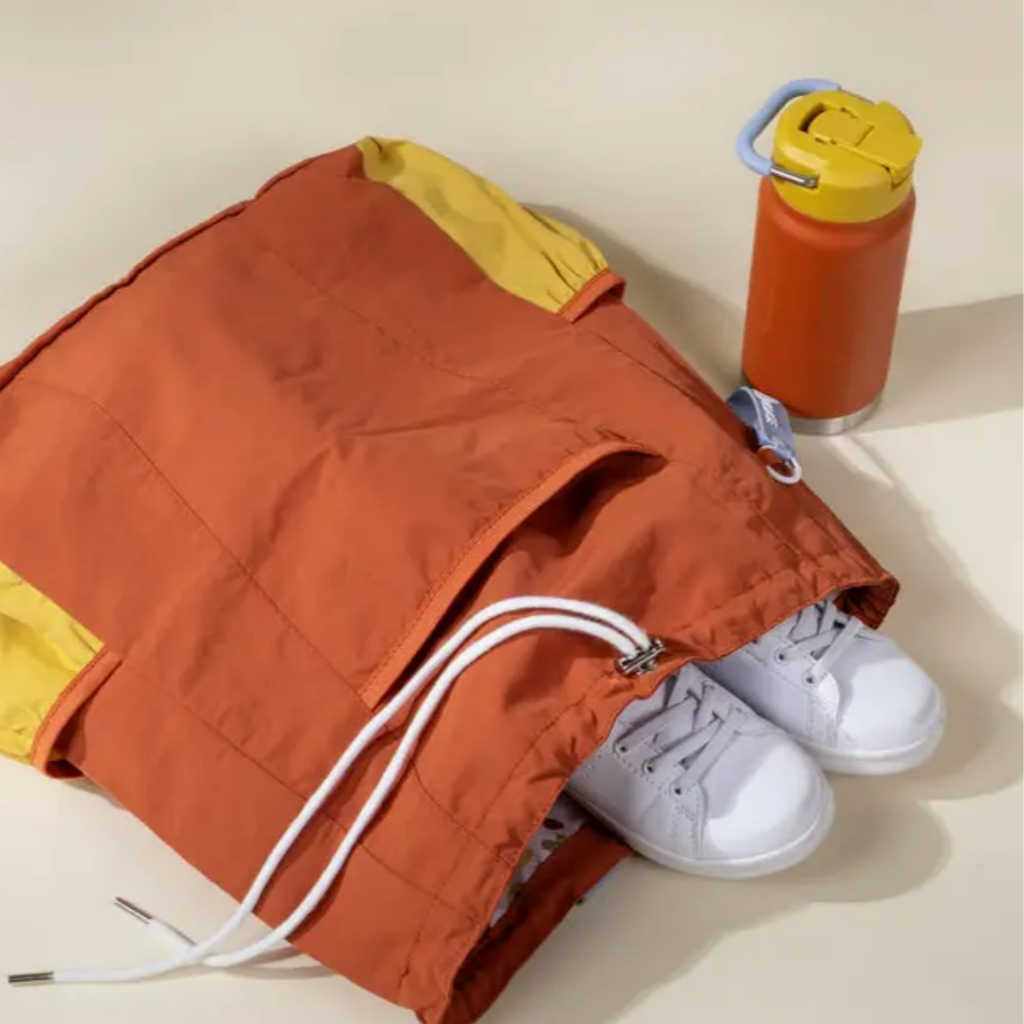 Orange drawstring bag with white sneakers and a red water bottle on a beige background