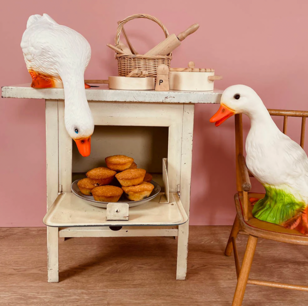 Decorative scene with goose figurines around a small oven and basket on a pink wall.