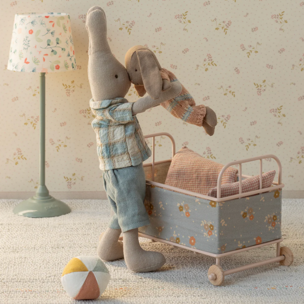 Stuffed bunny toy with a plaid shirt and shorts standing next to a small bed with pillows, in a room with floral wallpaper and a lamp.