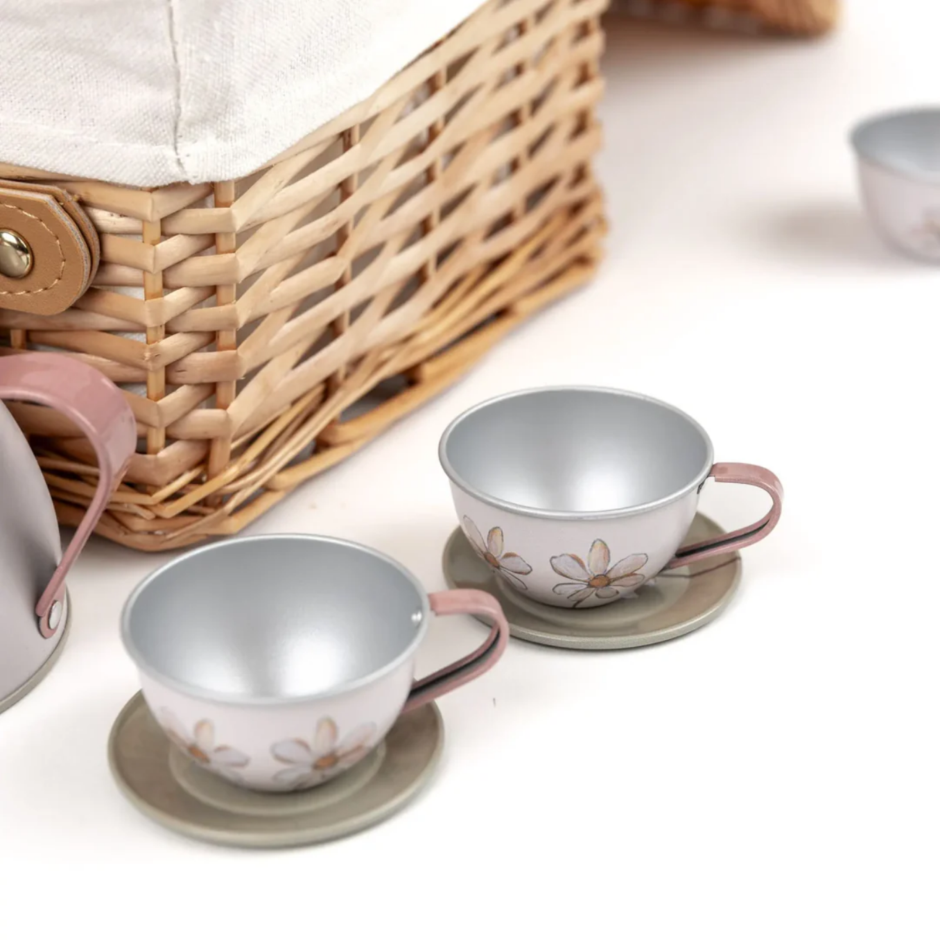 Two metal tea cups with floral designs on saucers next to a woven basket on a light surface.