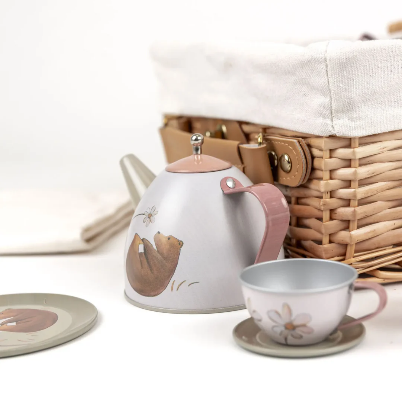 metal teapot and cup set with bear design in front of a wicker picnic basket.