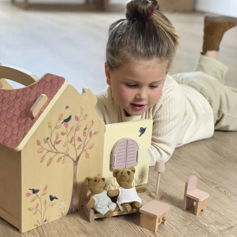 Child playing with a wooden toy house and teddy bears on a wooden floor.