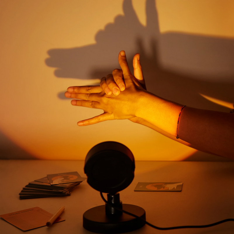 Hands reaching towards a black lamp on a table with cards, casting shadows on a yellow wall.
