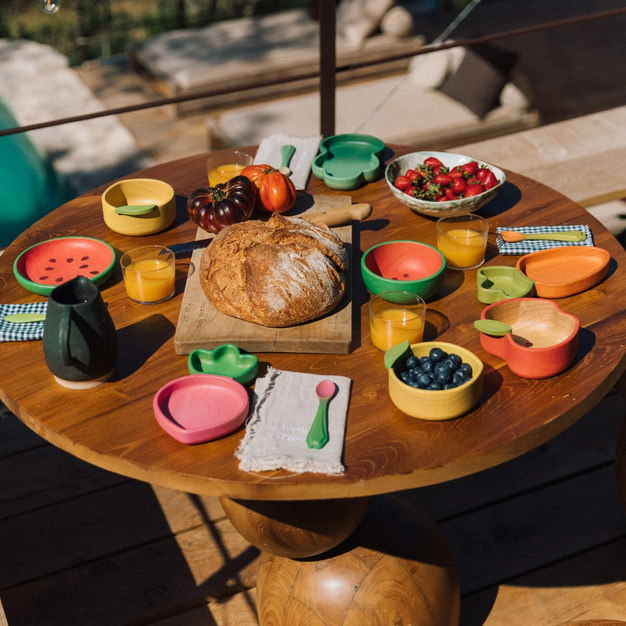 Wooden table with colorful ceramic plates, bowls, and a loaf of bread on an outdoor patio.